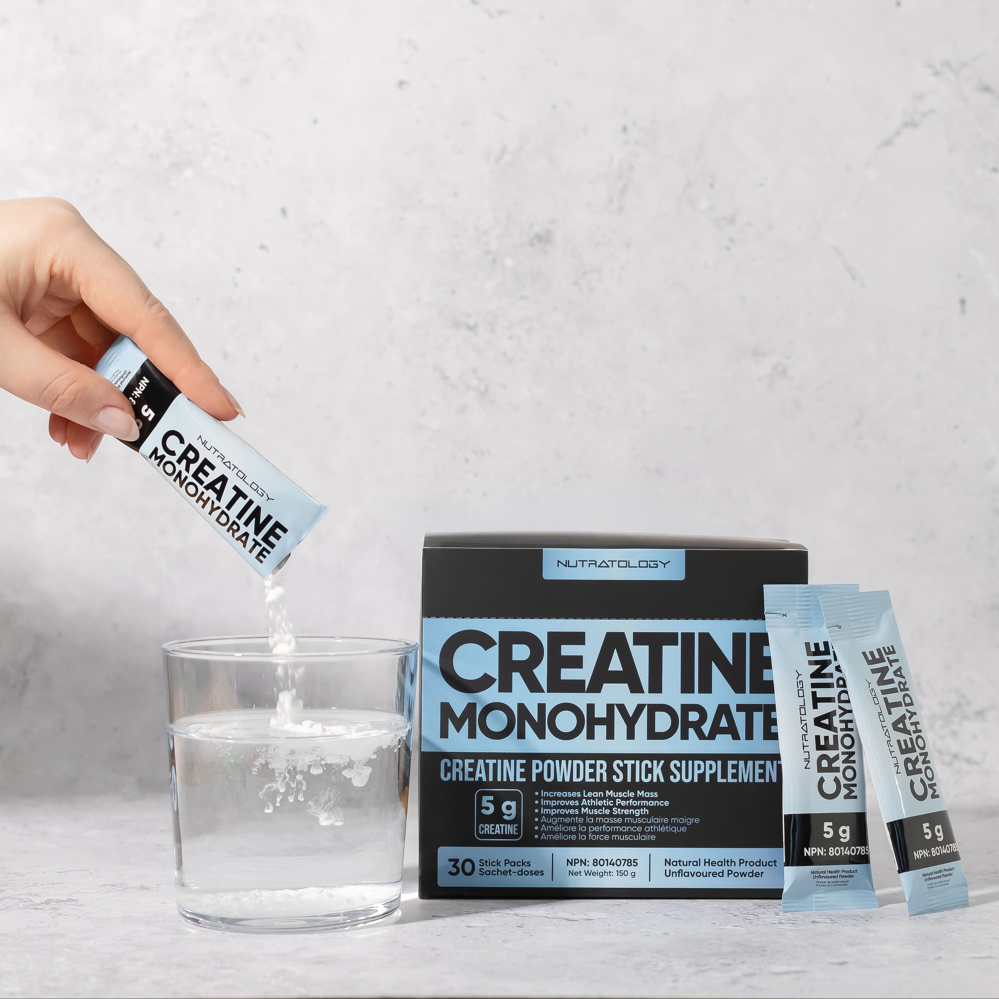 Person pouring creatine monohydrate powder from a stick into a glass of water, with packaging in the background.